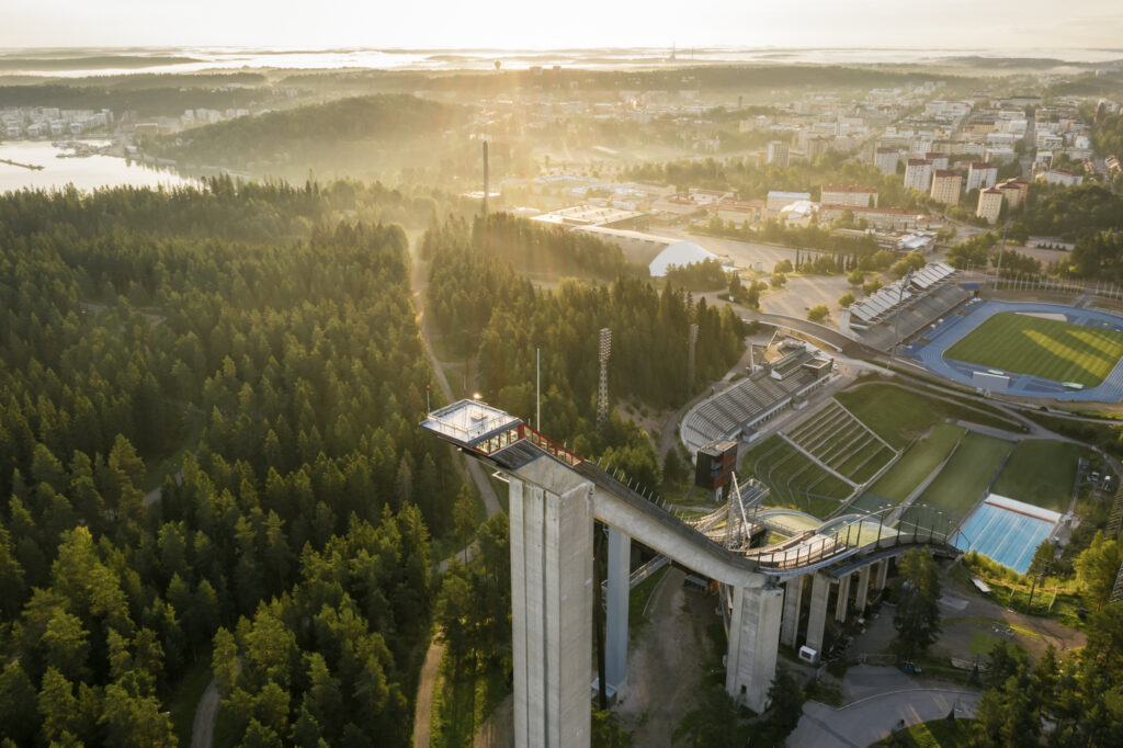 Ilmakuva, jossa etualalla hyppyrimäet ja taustalla auringonpaisteessa Lahden keskustaa.